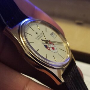 May include: A gold-toned wristwatch with a white face, featuring the text "HAMILTON MASTERPIECE" and a date display. The watch has a black leather band and a design with red, white, and blue accents. The watch is a classic timepiece.