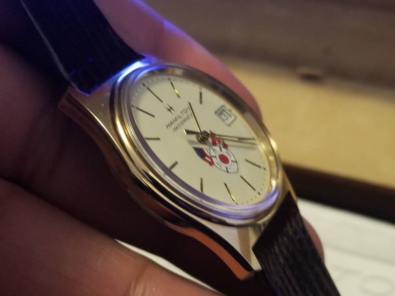 May include: A gold-toned wristwatch with a white face, featuring the text "HAMILTON MASTERPIECE" and a date display. The watch has a black leather band and a design with red, white, and blue accents. The watch is a classic timepiece.