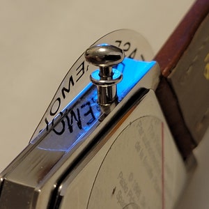 May include: A silver watch with a square face and a brown leather band. The watch has a silver knob on the side and a clear plastic cover over the face. The face of the watch has the words "EMOTION" printed on it.