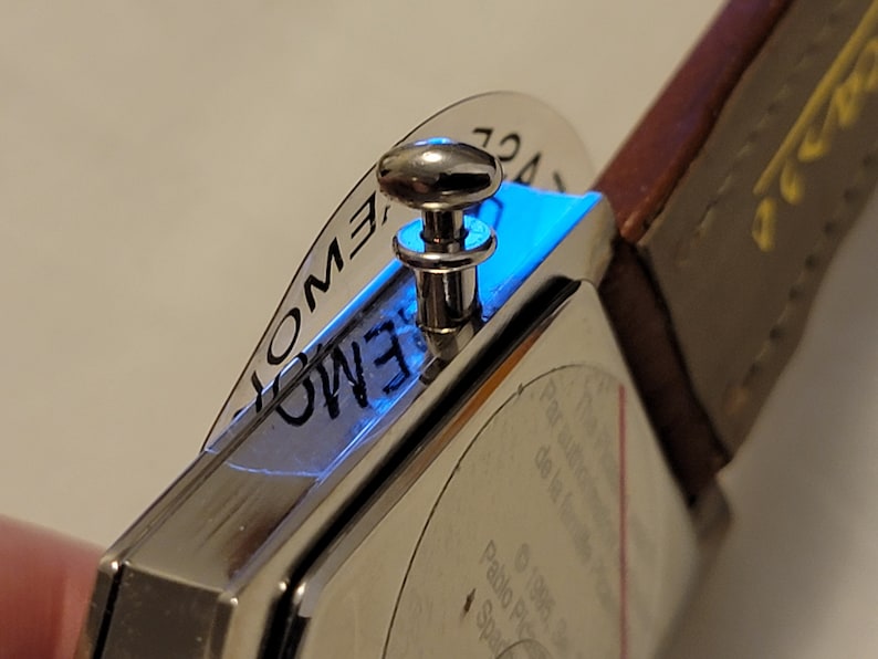 May include: A silver watch with a square face and a brown leather band. The watch has a silver knob on the side and a clear plastic cover over the face. The face of the watch has the words "EMOTION" printed on it.