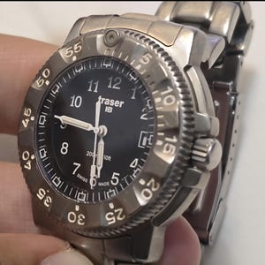 Puede incluir: Un reloj Traser H3 negro con una correa de metal plateado. La esfera del reloj tiene n&uacute;meros y manecillas blancos, y un bisel negro con n&uacute;meros blancos. El reloj es resistente al agua hasta 200 metros.