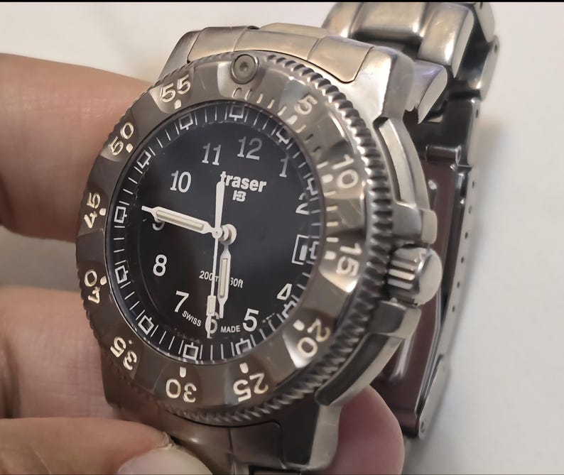 Puede incluir: Un reloj Traser H3 negro con una correa de metal plateado. La esfera del reloj tiene n&uacute;meros y manecillas blancos, y un bisel negro con n&uacute;meros blancos. El reloj es resistente al agua hasta 200 metros.