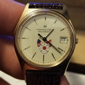 May include: A gold-toned Hamilton Masterpiece watch with a white face, featuring the IAFF logo and a date display. The watch has a brown leather band and is held by a hand.