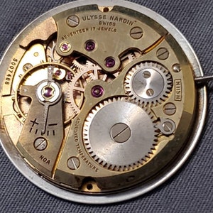 May include: Close-up of a gold-toned watch movement with the text "ULYSSE NARDIN" and "SWISS" on the top. The movement has a silver-toned center and is surrounded by a series of gears. The text "SEVENTEEN 17 JEWELS" is visible below the brand name. The text "ADJUSTED TEMPERATURE" is visible on the left side of the movement. The text "N115 H" is visible on the right side of the movement. The text "5007468" is visible on the left side of the movement.