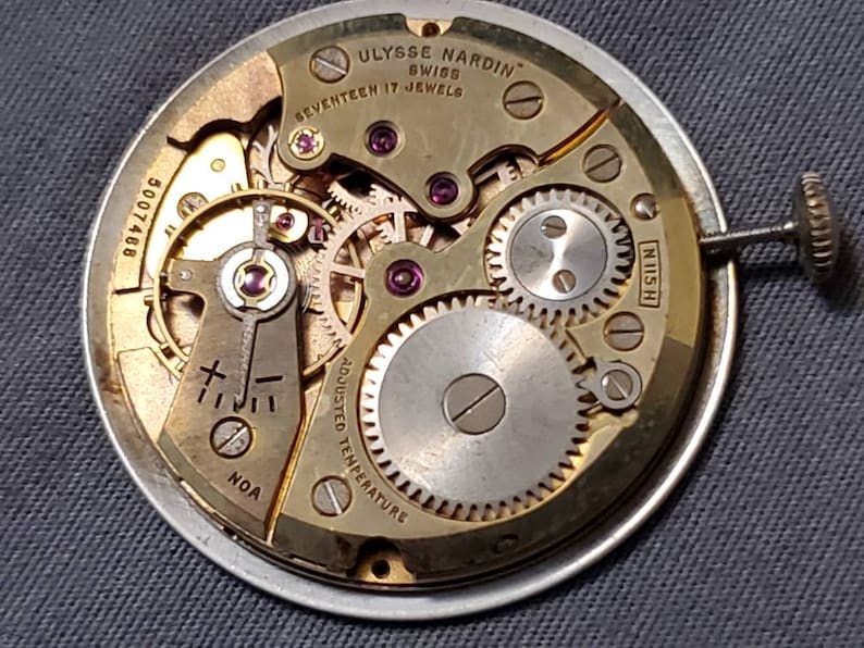 May include: Close-up of a gold-toned watch movement with the text "ULYSSE NARDIN" and "SWISS" on the top. The movement has a silver-toned center and is surrounded by a series of gears. The text "SEVENTEEN 17 JEWELS" is visible below the brand name. The text "ADJUSTED TEMPERATURE" is visible on the left side of the movement. The text "N115 H" is visible on the right side of the movement. The text "5007468" is visible on the left side of the movement.