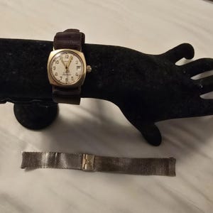 Könnte beinhalten: Eine goldfarbene Armbanduhr mit weißem Zifferblatt und schwarzem Lederband. Die Uhr befindet sich auf einem schwarzen Präsentationsständer. Ein silbernes Metallarmband ist ebenfalls abgebildet.