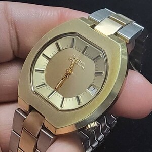 K&ouml;nnte beinhalten: Eine goldfarbene Armbanduhr mit quadratischem Zifferblatt und einem zweifarbigen Metallband. Das Zifferblatt hat einen goldenen Hintergrund mit schwarzen Stunden- und Minutenzeigern und einem wei&szlig;en Datumsfenster bei 3 Uhr. Auf dem Zifferblatt ist auch der Text "Chronograph" aufgedruckt.