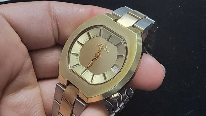 K&ouml;nnte beinhalten: Eine goldfarbene Armbanduhr mit quadratischem Zifferblatt und einem zweifarbigen Metallband. Das Zifferblatt hat einen goldenen Hintergrund mit schwarzen Stunden- und Minutenzeigern und einem wei&szlig;en Datumsfenster bei 3 Uhr. Auf dem Zifferblatt ist auch der Text "Chronograph" aufgedruckt.