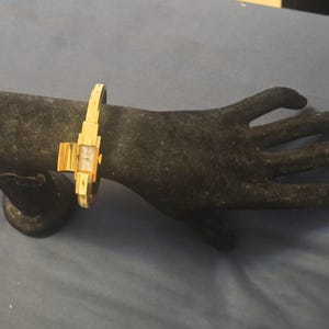 May include: A gold-tone bangle watch featuring a rectangular white face with thin black hands and markers. The watch band is a rigid, hinged metal cuff design with a polished finish, showcasing a sleek, streamlined aesthetic for the wrist.