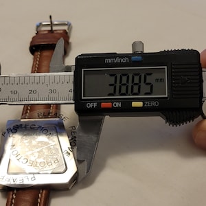 May include: A silver watch face with a protective film that reads "Please remove protection". The watch face is being measured with a digital caliper that reads 38.85 mm.