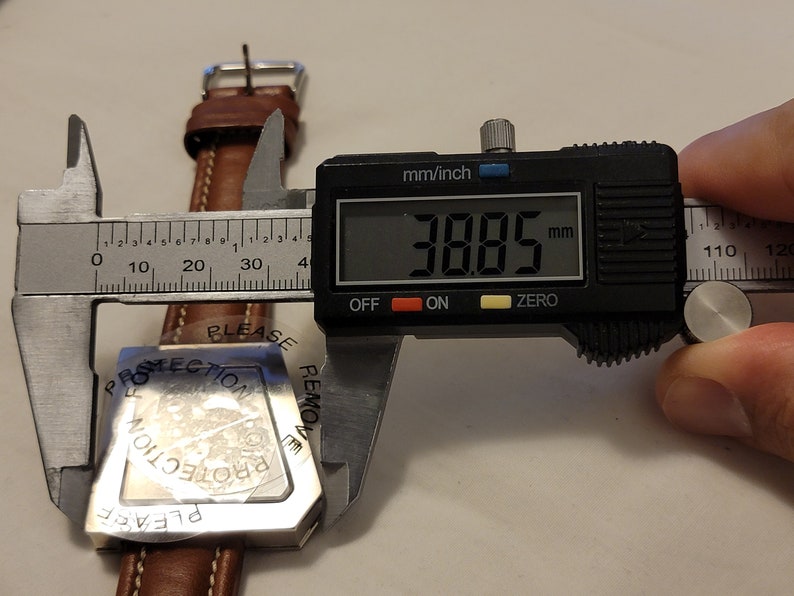 May include: A silver watch face with a protective film that reads "Please remove protection". The watch face is being measured with a digital caliper that reads 38.85 mm.