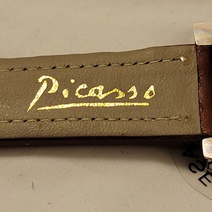 May include: A brown leather watch band with gold stitching and the word "Picasso" written in gold lettering.