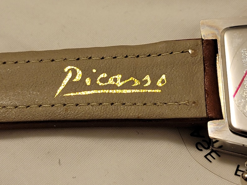 May include: A brown leather watch band with gold stitching and the word "Picasso" written in gold lettering.