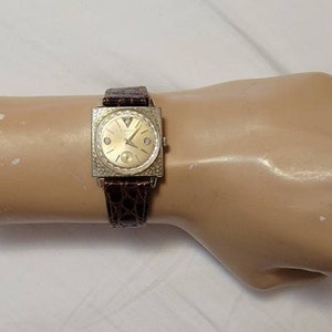 Op de afbeelding: Een vierkante gouden polshorloge met een bruin leren bandje. De wijzerplaat van het horloge heeft een witte achtergrond met zwarte uur- en minutenwijzers en zwarte Romeinse cijfers als uurmarkeringen.