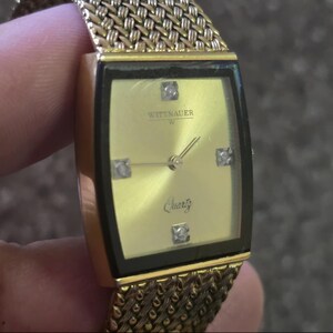 May include: A gold-tone Wittnauer watch with a square face and a gold mesh band. The watch face is white with black Roman numeral hour markers and black hands. The watch face also has a small diamond at the 12 o'clock position and the word "Quartz" printed in black.