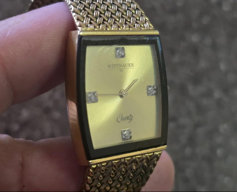May include: A gold-tone Wittnauer watch with a square face and a gold mesh band. The watch face is white with black Roman numeral hour markers and black hands. The watch face also has a small diamond at the 12 o'clock position and the word "Quartz" printed in black.