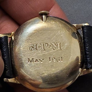 May include: A gold-toned wristwatch with a black leather band. The back of the watch has the inscription "J.P.M. May 1961".