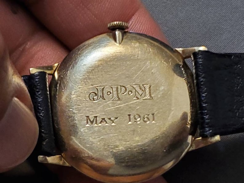 May include: A gold-toned wristwatch with a black leather band. The back of the watch has the inscription "J.P.M. May 1961".