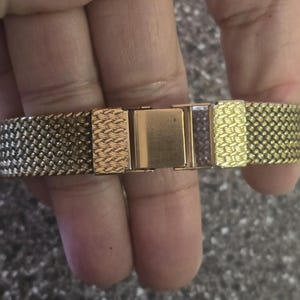 May include: A gold-tone watch band with a woven mesh design. The band has a clasp closure.