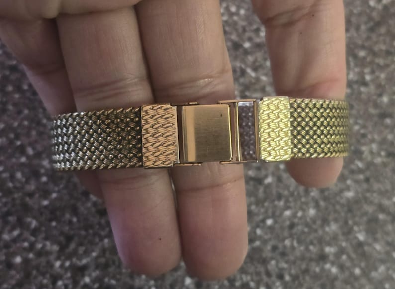 May include: A gold-tone watch band with a woven mesh design. The band has a clasp closure.