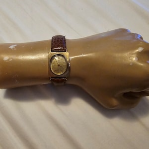 Op de afbeelding: Een gouden polshorloge met een rechthoekige wijzerplaat en een bruin leren bandje. Het horloge zit om de pols van een mannequin.