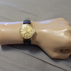 Puede incluir: Un reloj de pulsera dorado con una correa de cuero marrón. La esfera del reloj es redonda y tiene un fondo blanco con marcas negras. El reloj tiene una ventana de fecha en la posición de las 3 en punto.