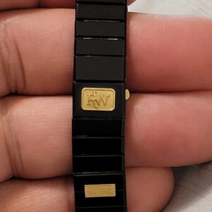 May include: A black and gold bracelet with a gold 'RW' logo. The bracelet is made of linked metal pieces.