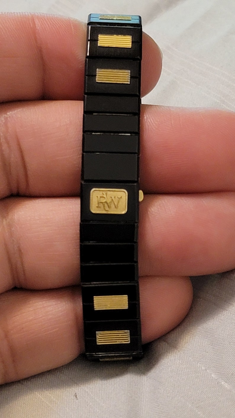 May include: A black and gold bracelet with a gold 'RW' logo. The bracelet is made of linked metal pieces.