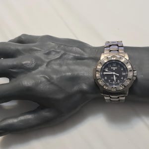 Puede incluir: Un reloj de pulsera plateado con una esfera negra y una ventana de fecha. El reloj tiene un bisel negro con marcas blancas y una correa plateada.