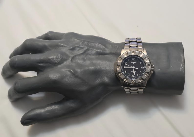 Puede incluir: Un reloj de pulsera plateado con una esfera negra y una ventana de fecha. El reloj tiene un bisel negro con marcas blancas y una correa plateada.