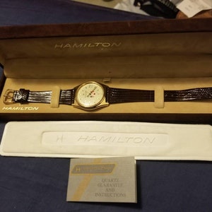 May include: A gold-toned Hamilton watch with a brown leather band, displayed in a brown velvet box. The watch has a cream-colored face with gold accents and a second hand. The box is open, revealing the watch and a white insert. A warranty card is also present.
