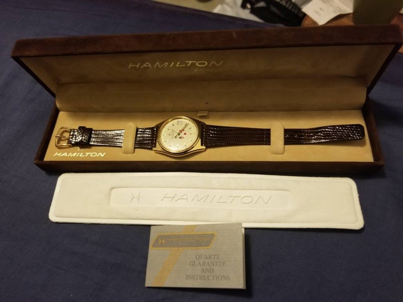 May include: A gold-toned Hamilton watch with a brown leather band, displayed in a brown velvet box. The watch has a cream-colored face with gold accents and a second hand. The box is open, revealing the watch and a white insert. A warranty card is also present.