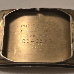 以下が含まれることがあります： 銀色の腕時計の裏側のクローズアップ。刻印は「Cased & Timed in U.S.A. by The Gruen Watch Co. 370/716 C346525」です。