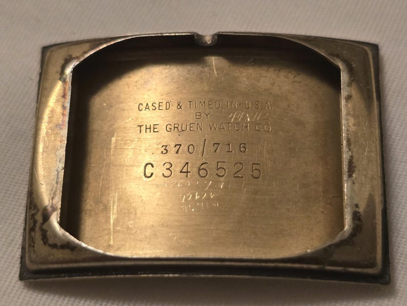 以下が含まれることがあります： 銀色の腕時計の裏側のクローズアップ。刻印は「Cased & Timed in U.S.A. by The Gruen Watch Co. 370/716 C346525」です。