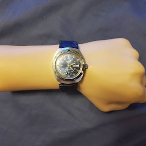 Peut inclure: Une montre-bracelet argentée et noire avec un cadran bleu et des accents dorés. La montre a un guichet date et une lunette tournante.