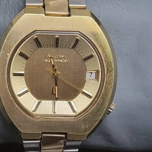 K&ouml;nnte beinhalten: Eine goldfarbene Armbanduhr mit einem runden Zifferblatt und einem silbernen Armband. Das Zifferblatt hat einen goldenen Hintergrund mit schwarzen Markierungen und einem wei&szlig;en Datumsfenster. Auf dem Zifferblatt steht auch der Text "Bulova Accutron".