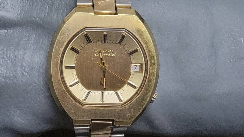 K&ouml;nnte beinhalten: Eine goldfarbene Armbanduhr mit einem runden Zifferblatt und einem silbernen Armband. Das Zifferblatt hat einen goldenen Hintergrund mit schwarzen Markierungen und einem wei&szlig;en Datumsfenster. Auf dem Zifferblatt steht auch der Text "Bulova Accutron".