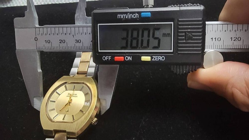 K&ouml;nnte beinhalten: Eine goldfarbene Uhr mit quadratischem Zifferblatt und einem Metallband. Die Uhr wird mit einer Schieblehre gemessen, die 38,05 mm anzeigt.