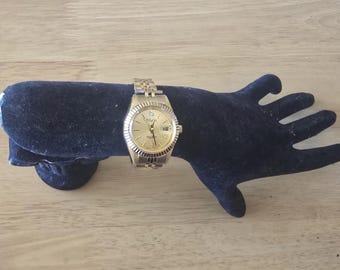 May include: A gold-tone wristwatch with a white face and black Roman numeral hour markers. The watch has a diamond at the 12 o'clock position and a date window at the 3 o'clock position.