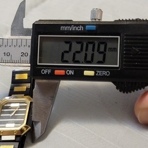 May include: A black and gold watch with a square face is being measured with a digital caliper. The caliper reads 22.09 mm.