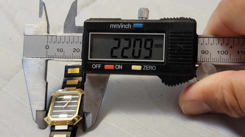 May include: A black and gold watch with a square face is being measured with a digital caliper. The caliper reads 22.09 mm.