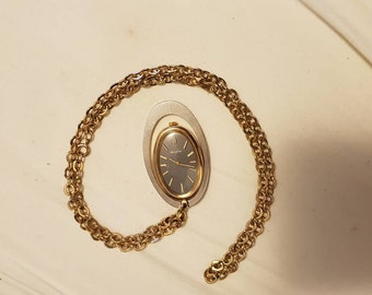 Könnte beinhalten: Eine goldene Kettenkette mit einem ovalen Uhrenanhänger. Das Zifferblatt der Uhr ist silberfarben mit schwarzen Zeigern und schwarzen römischen Ziffern. Auf dem Zifferblatt der Uhr steht "Bulova" gedruckt.