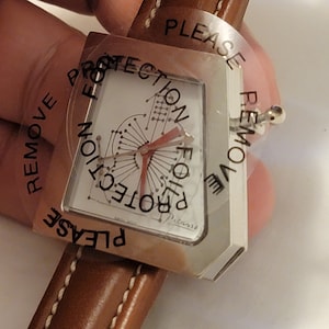 May include: A silver watch with a white face and a brown leather band. The watch face has a unique design with a red hand and the text "PLEASE REMOVE PROTECTION FOR PROTECTION" printed on the clear plastic cover.