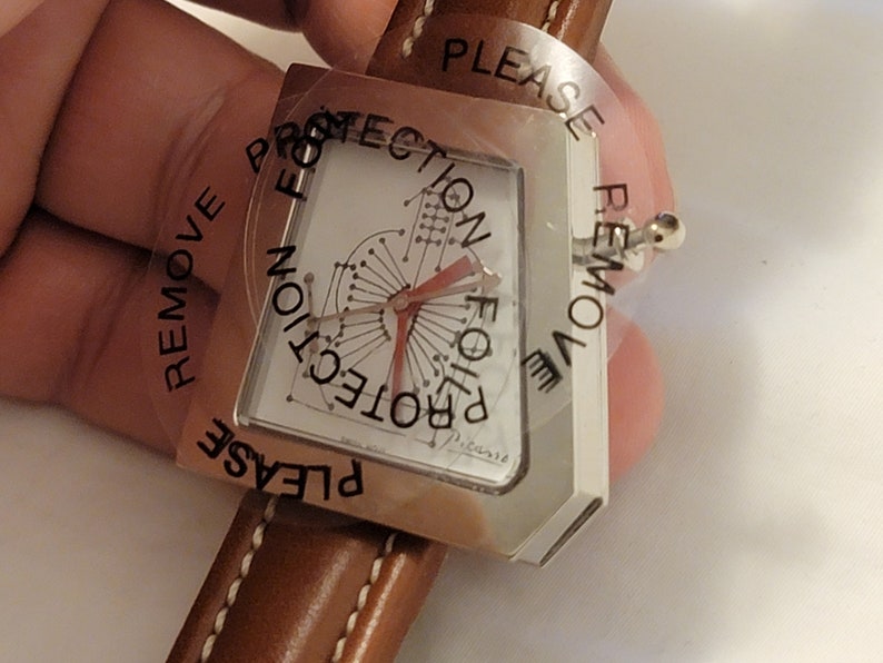 May include: A silver watch with a white face and a brown leather band. The watch face has a unique design with a red hand and the text "PLEASE REMOVE PROTECTION FOR PROTECTION" printed on the clear plastic cover.