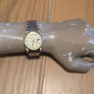 Op de afbeelding: Een tweekleurig polshorloge met een goudkleurige gekartelde rand en een crèmekleurige wijzerplaat met een dag-datumvenster op de twaalf- en drie-uurpositie. Het horloge heeft een metalen schakelarmband met afwisselende zilveren en gouden tinten.