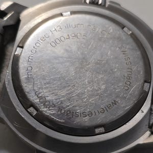 Puede incluir: Primer plano de la parte posterior de un reloj plateado con el texto "mb-microtec H3 illum" y "0004905" grabado en la caja. El reloj tambi&eacute;n tiene el texto "Water resist" y "made swiss" grabado en la caja.