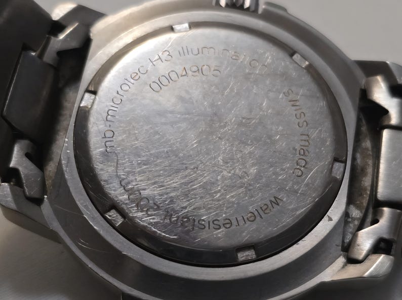Puede incluir: Primer plano de la parte posterior de un reloj plateado con el texto "mb-microtec H3 illum" y "0004905" grabado en la caja. El reloj tambi&eacute;n tiene el texto "Water resist" y "made swiss" grabado en la caja.