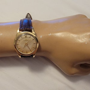 Puede incluir: Un reloj de pulsera de oro con una esfera blanca y una correa de cuero negra en una muñeca de maniquí.