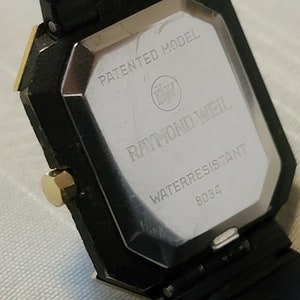May include: The back of a black and gold Raymond Weil watch with a silver face. The watch is water resistant and has a patented model number. The watch face says "Raymond Weil" and "Water Resistant 8034".