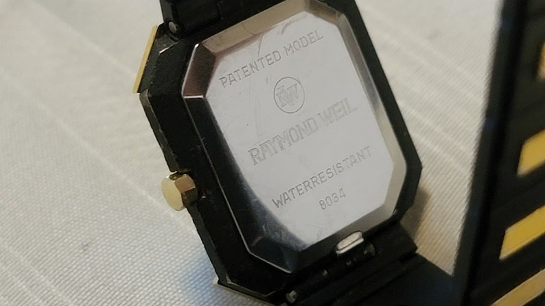 May include: The back of a black and gold Raymond Weil watch with a silver face. The watch is water resistant and has a patented model number. The watch face says "Raymond Weil" and "Water Resistant 8034".